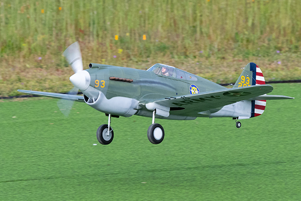 Model airplane with green body and spinning propeller flying over grassy field.