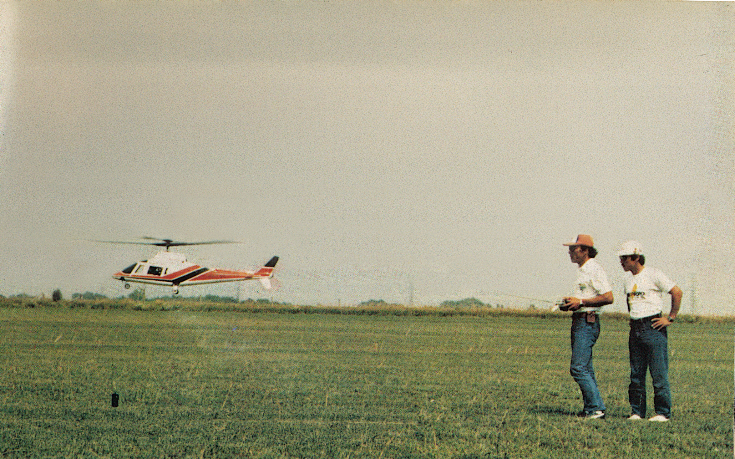 A remote-controlled helicopter flies low; two men watch, standing on grass.