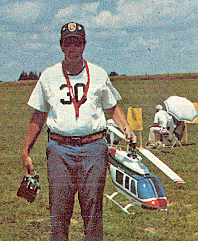 Man holding remote with large model helicopter outdoors, wearing a cap and sunglasses.