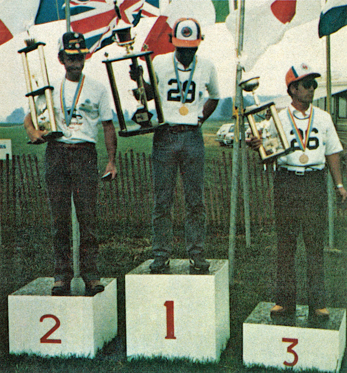 Three men on a podium holding trophies, wearing medals and caps, numbered 1, 2, and 3.