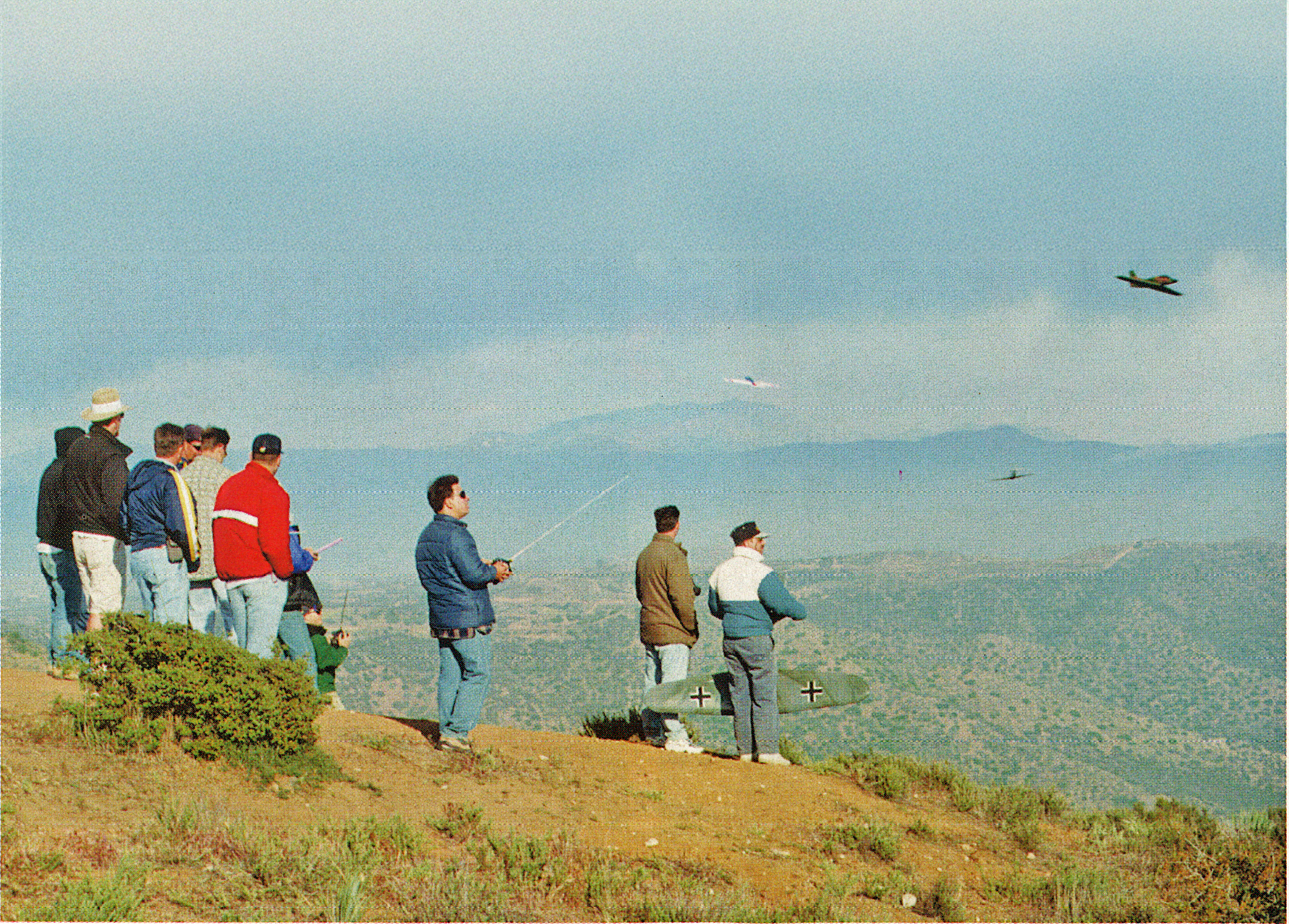 Group flying remote-controlled planes on a hilltop with a scenic view.