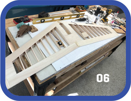 Wooden model plane wing on a workbench, tools scattered, number "06" in white.
