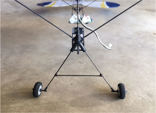 Rear view of a small model airplane with exposed wires and landing gear on a concrete floor.