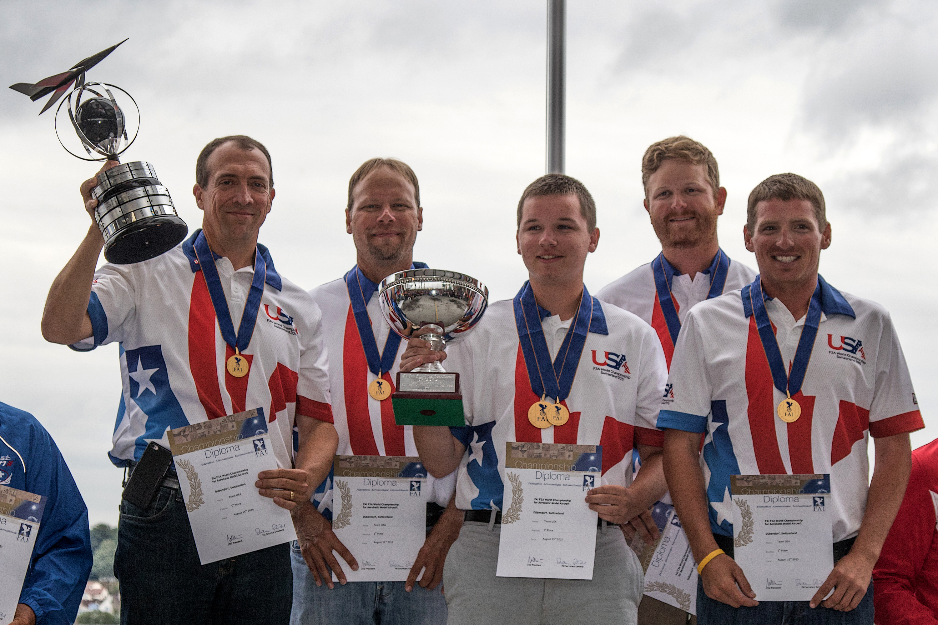 Five men in matching USA shirts hold medals, trophies, and certificates, smiling.