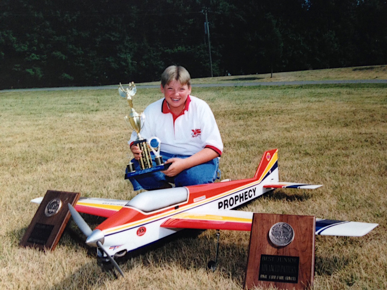 Young person with model airplane and trophies on grass.