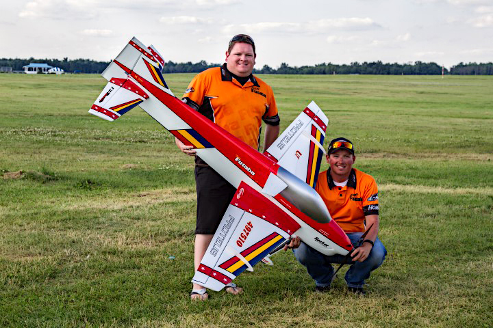 Two people with a large model airplane on a grassy field. Both wear orange shirts.