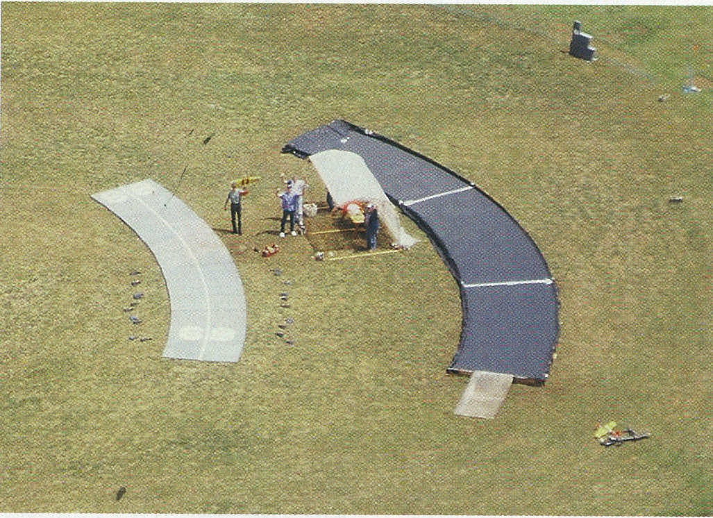 Curved structures on grass with people assembling in the center.