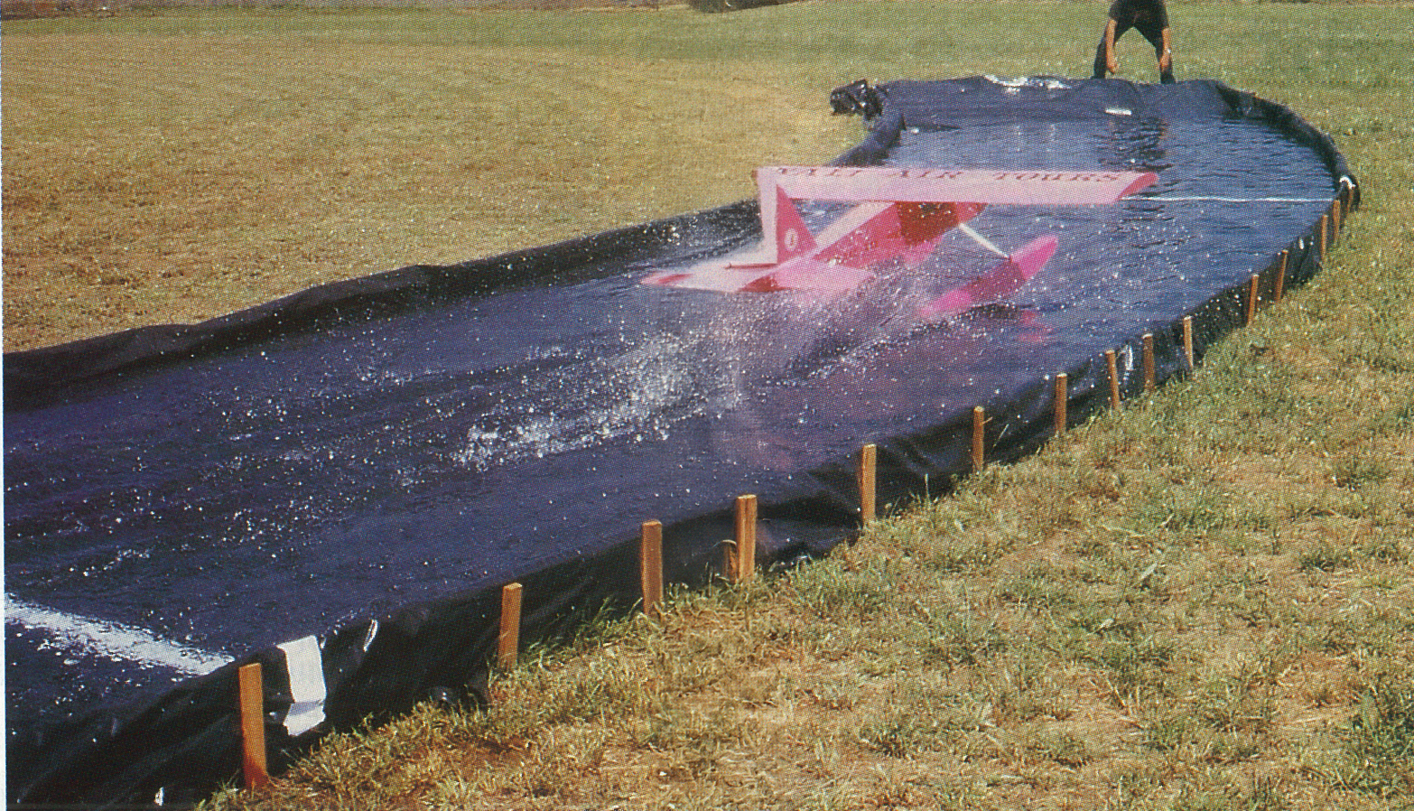 Pink toy plane skids across a water slide on a grassy field.