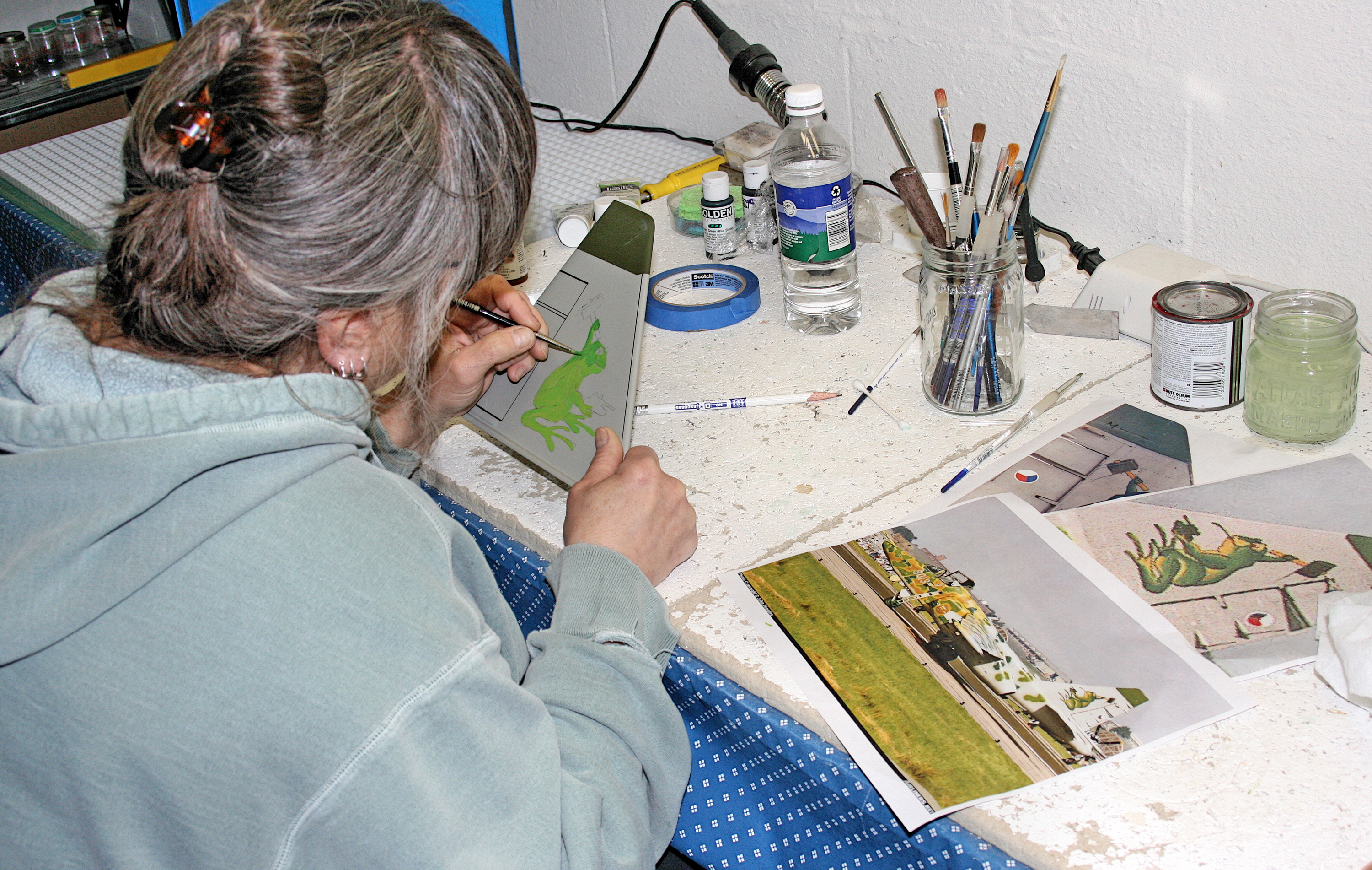Artist painting at a cluttered desk with brushes, jars, and photo references.