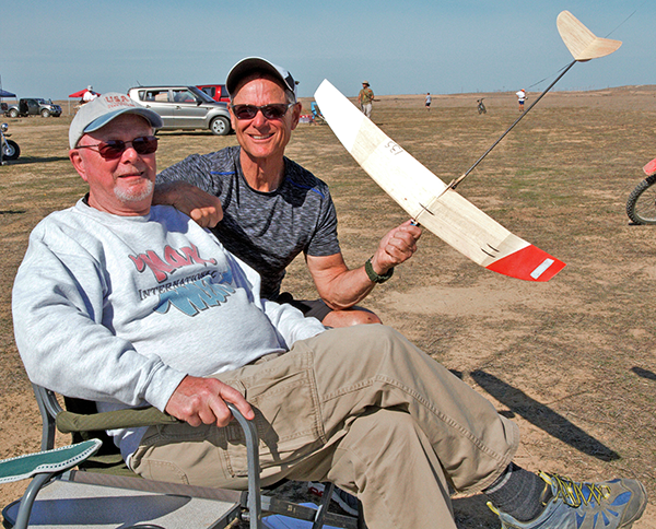 Two men smiling outdoors. One seated, the other holding a model glider.