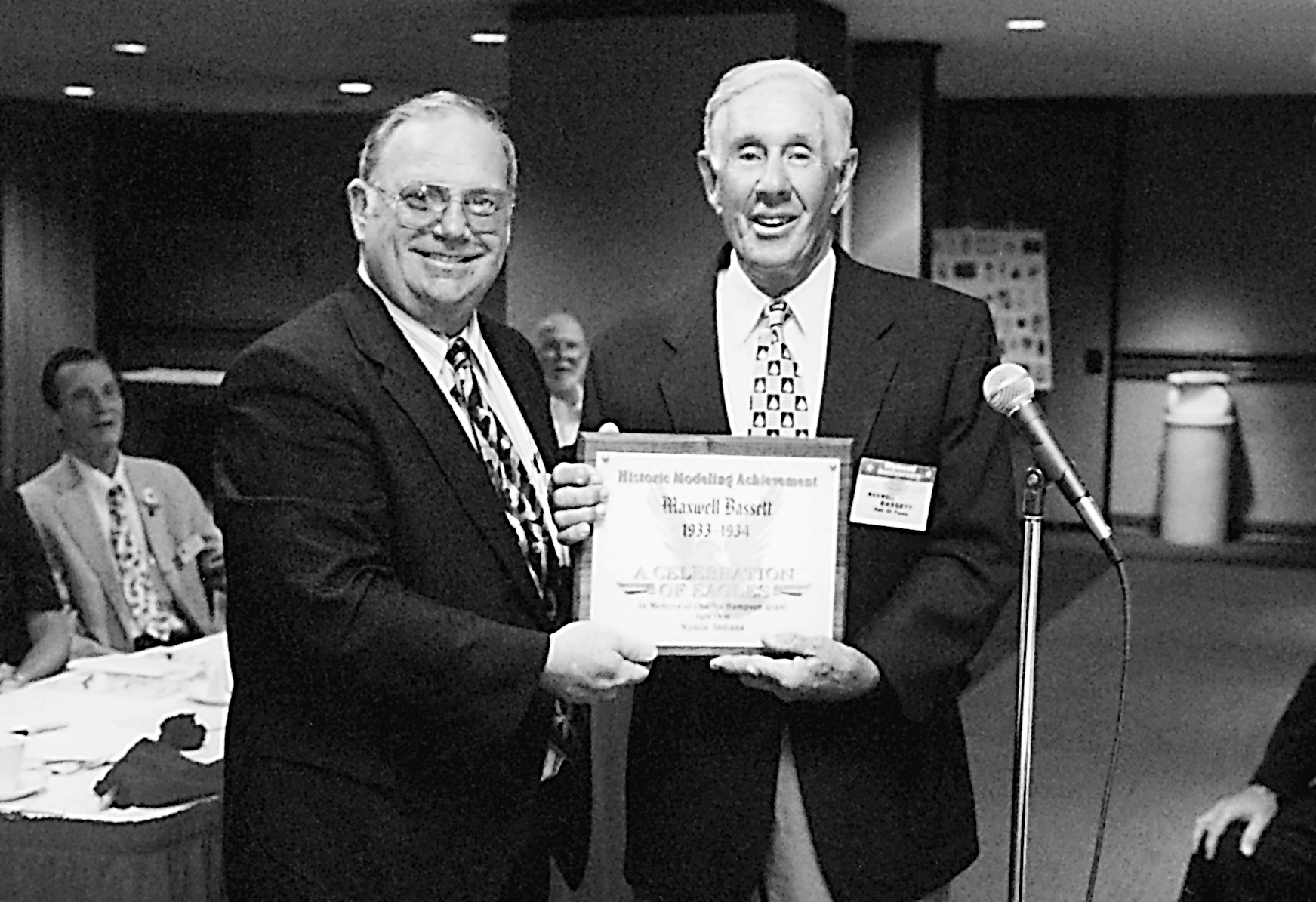 Two men smiling, holding a framed certificate at an indoor event.