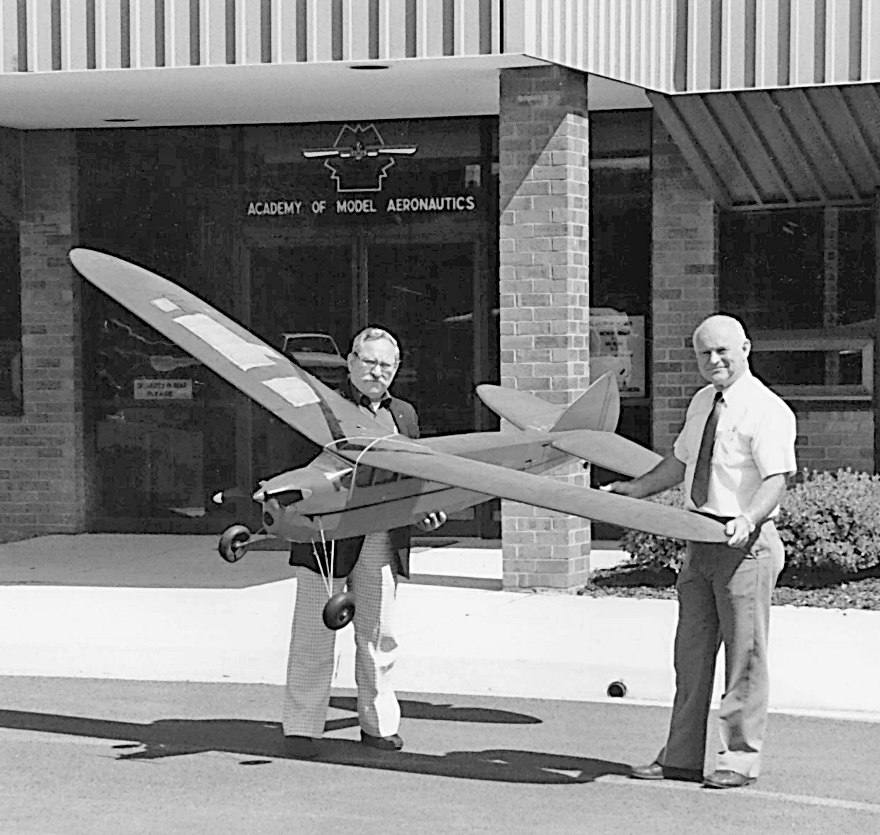 Two men holding a large model airplane outside a building labeled "Academy of Model Aeronautics."