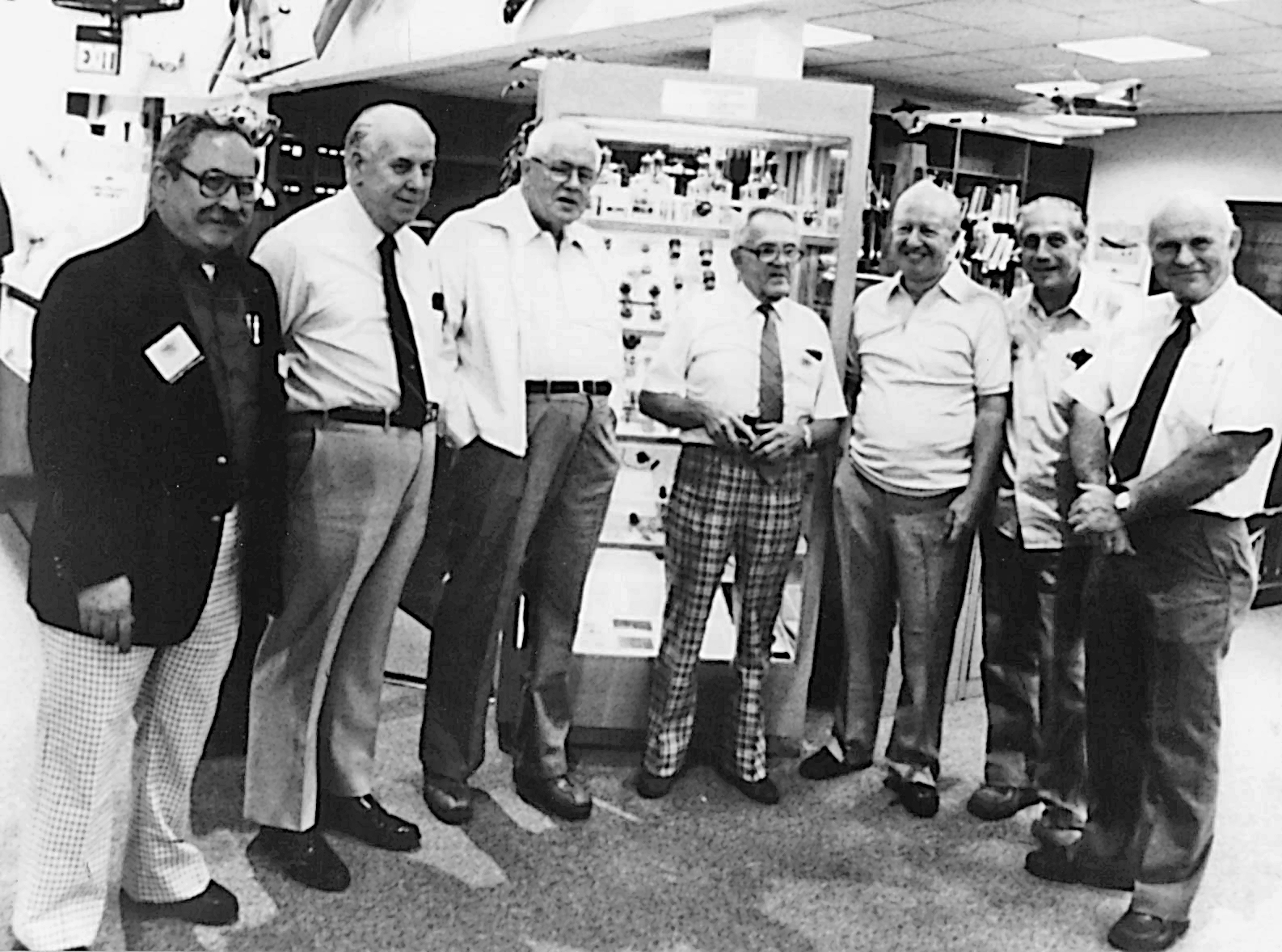Seven men stand together, smiling, in a library or bookstore.