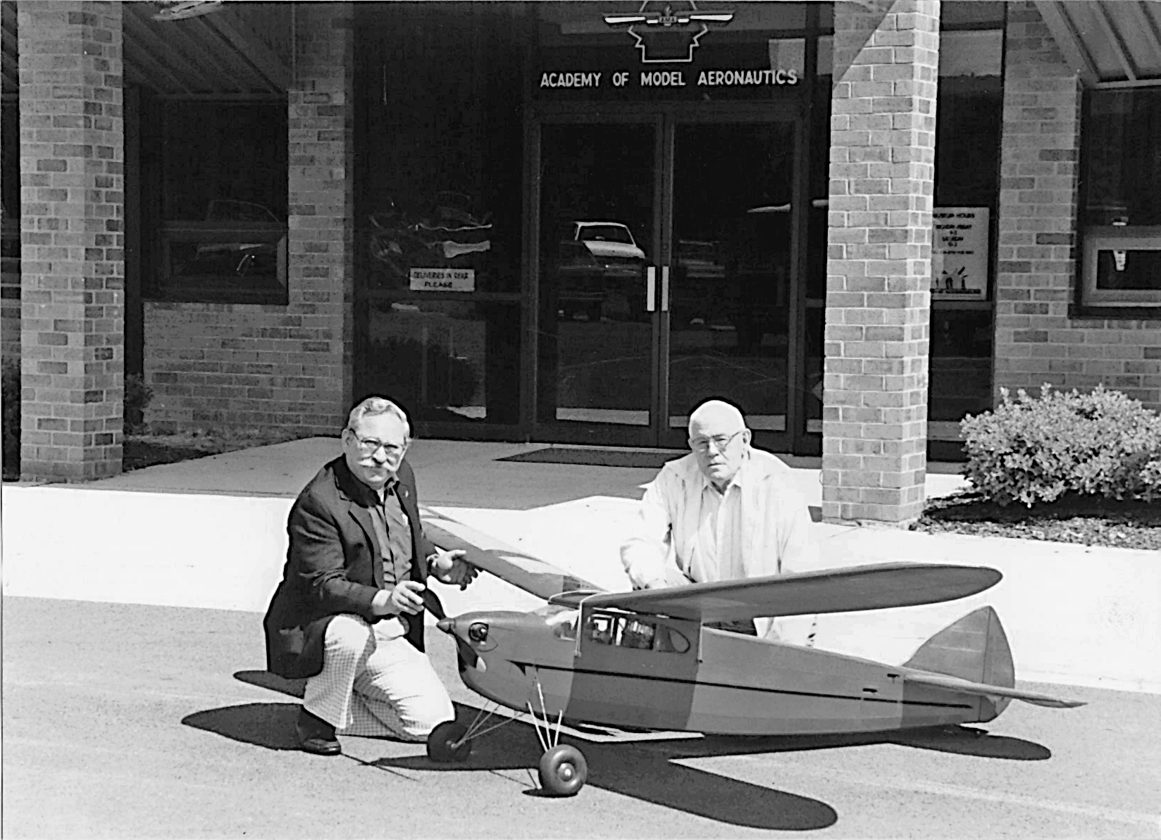 Two men kneeling by model airplane outside brick building labeled "Academy of Model Aeronautics."