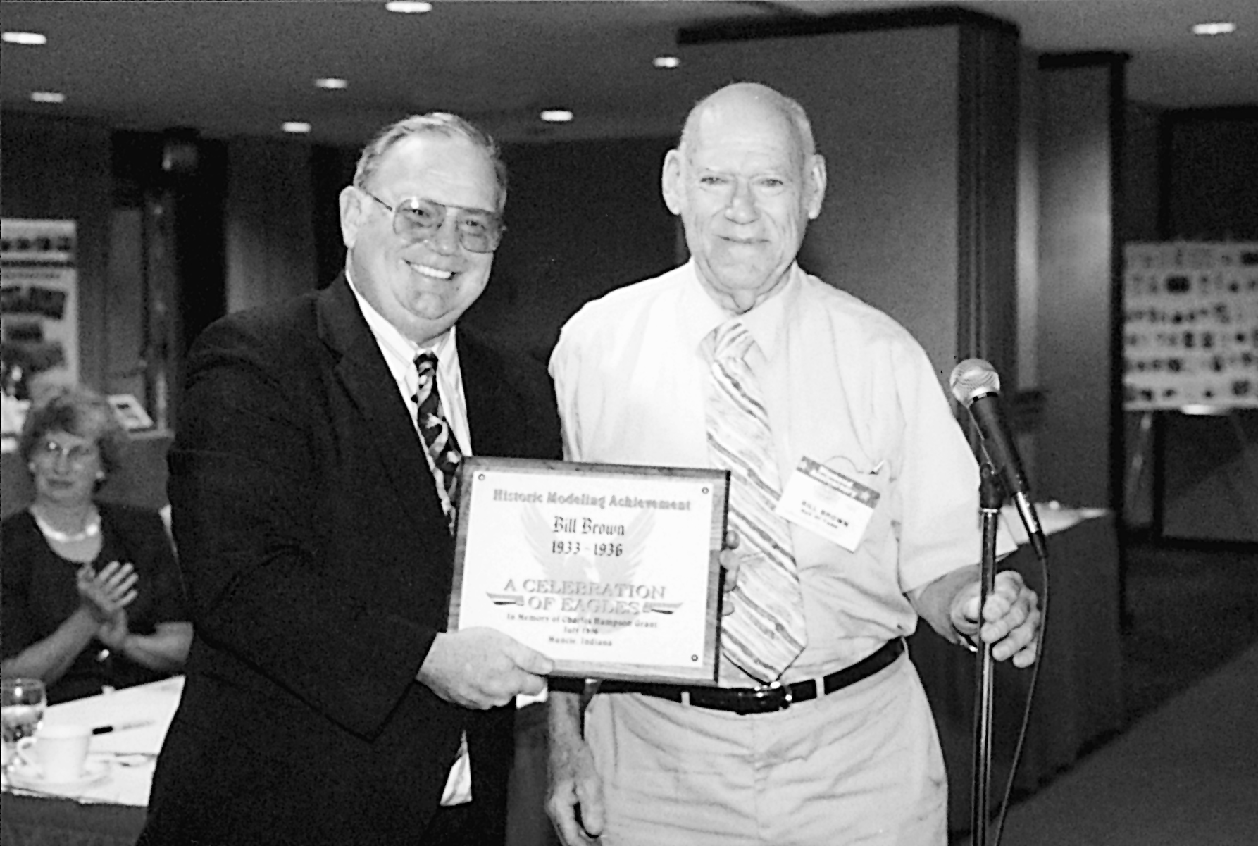 Two men in a room, one holding an award plaque, smiling, in black and white.