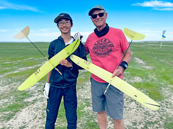 Two people smiling, holding yellow model gliders on a grassy field.