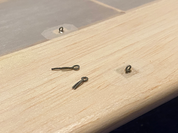 Small metal hook screws on a wooden board, some taped down.