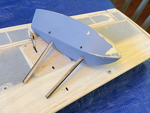 Model boat hull on a wooden base with metal supports, blue tarp background.