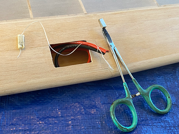 Wooden model wing with wires and a small electronic component next to green-handled pliers.