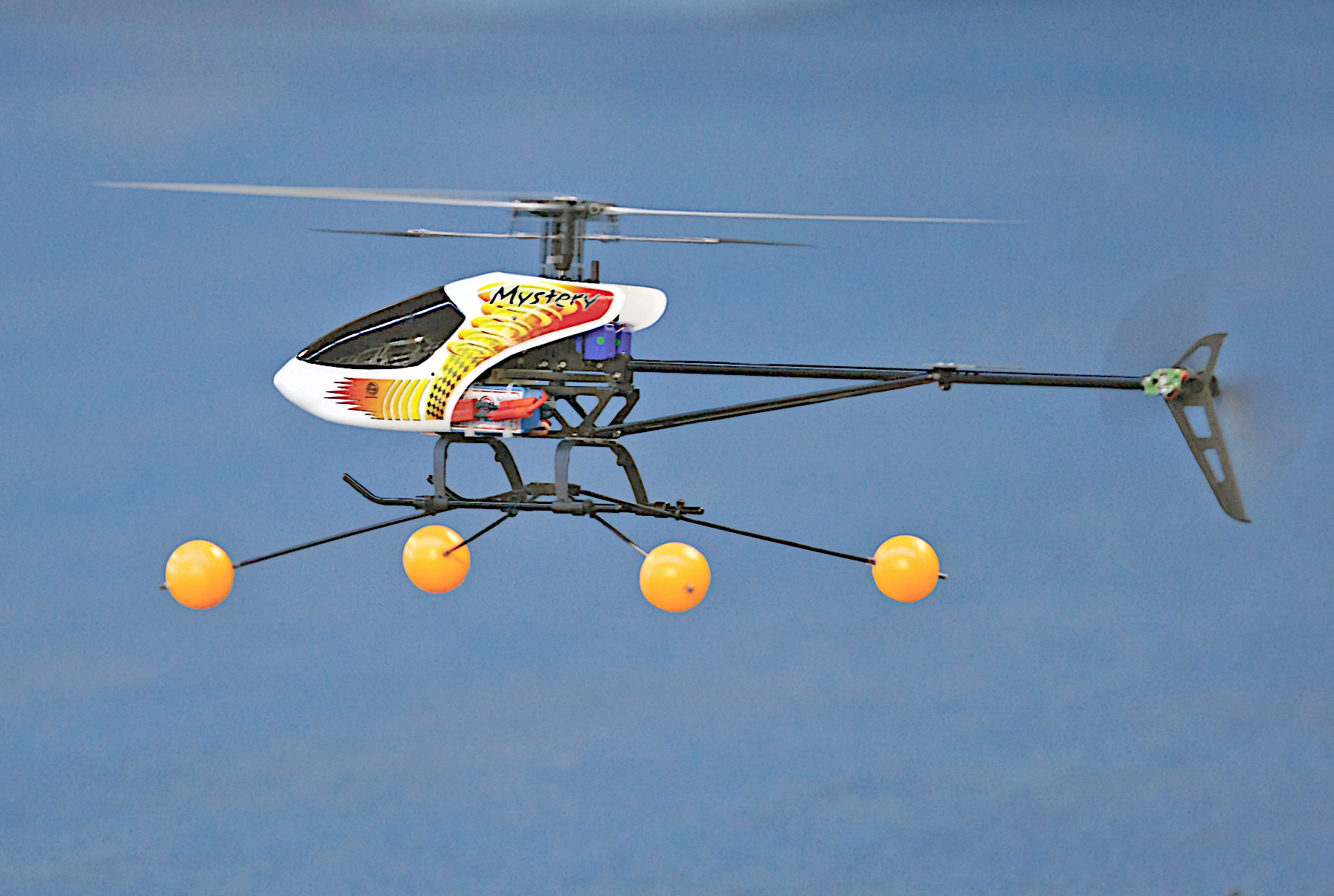 Toy helicopter flying against a blue sky, with yellow balls on landing gear.