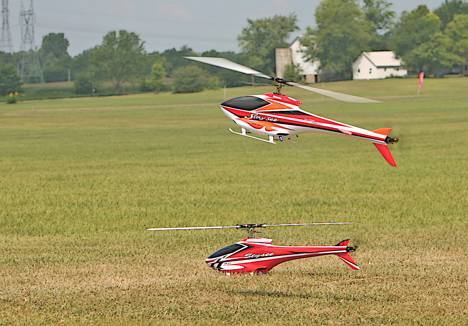 Two red remote-controlled helicopters flying over a grassy field.
