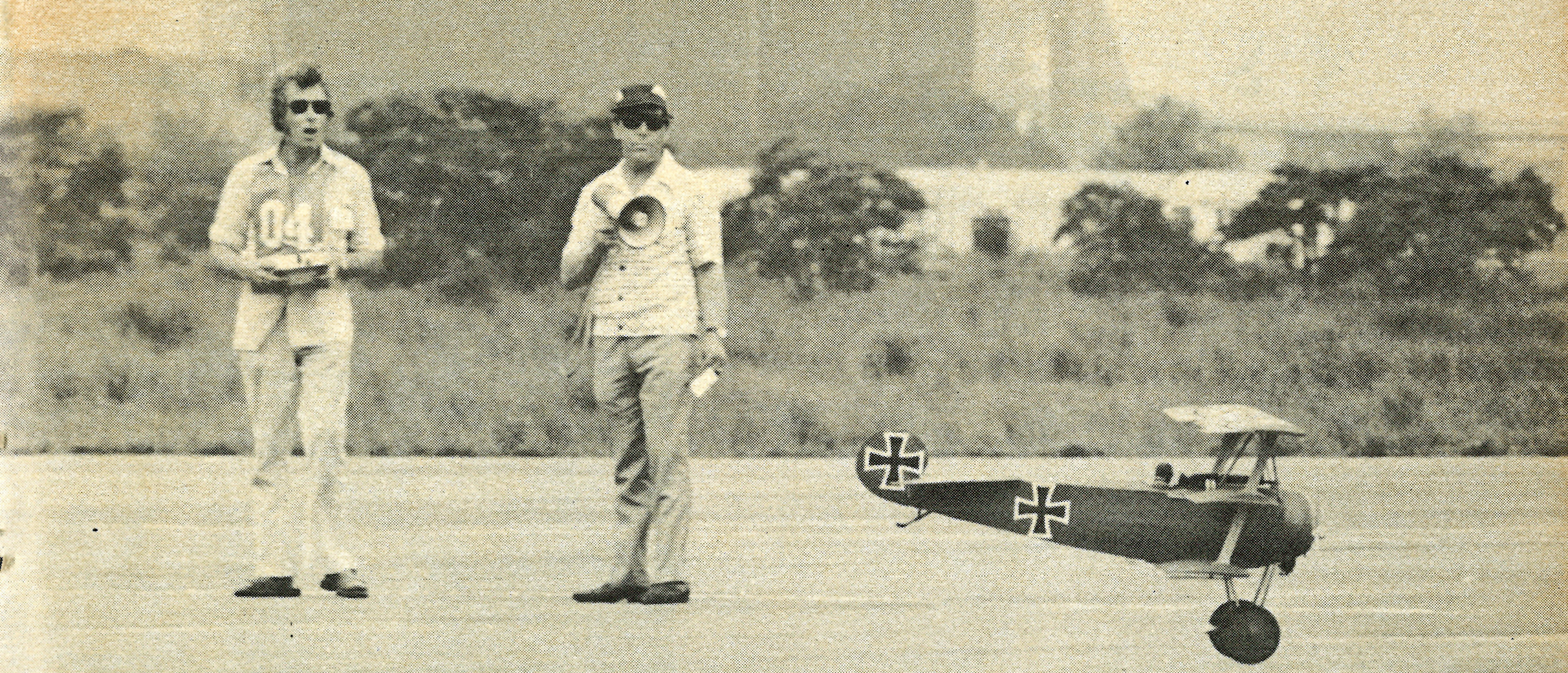 Two men stand on a runway with a model plane.