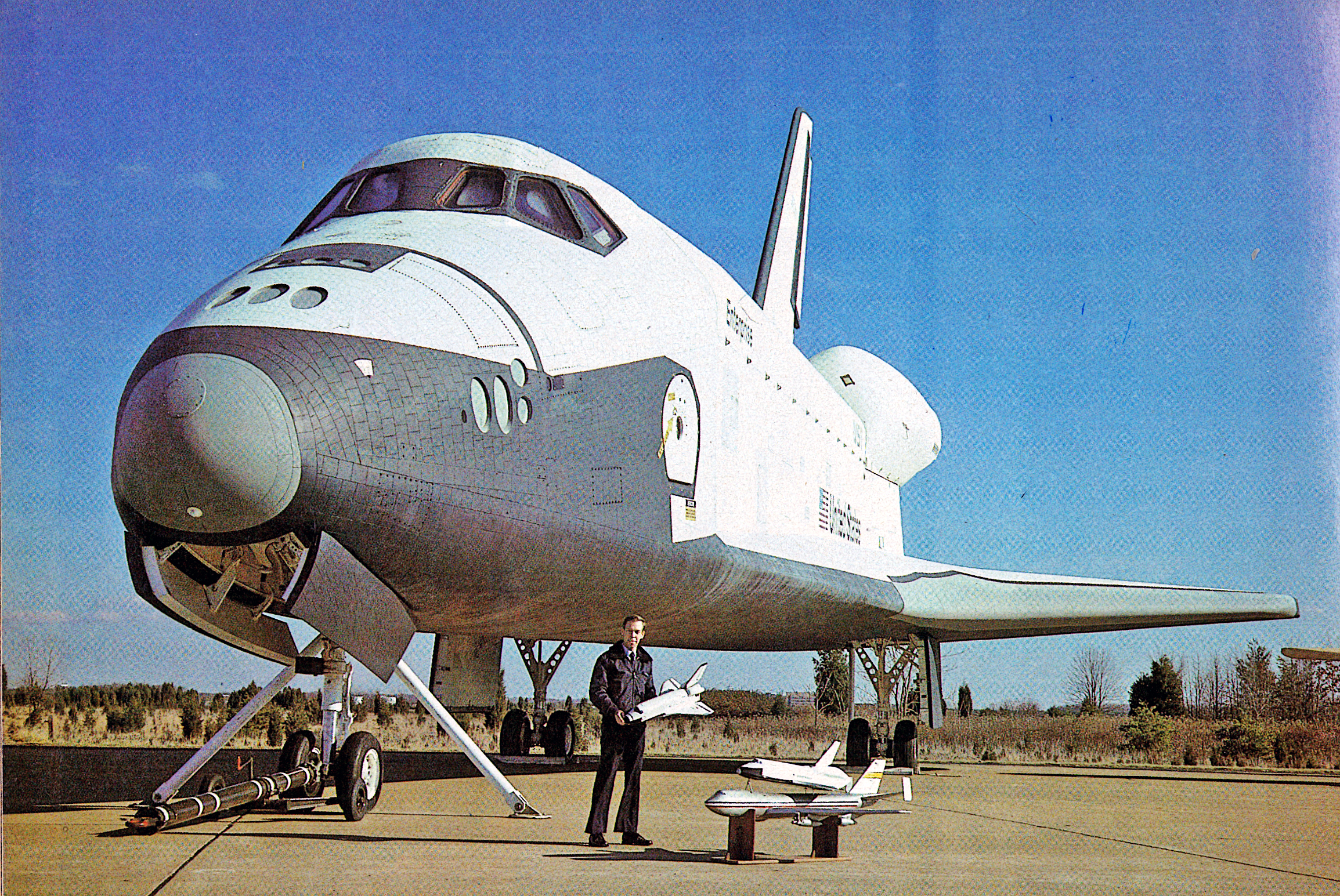 Space shuttle on runway, person holding model shuttle, clear blue sky.