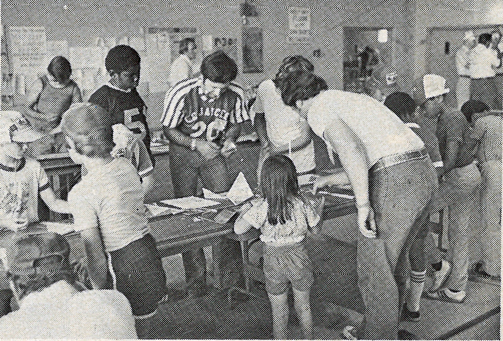 People gathered around tables, interacting and filling out forms in a busy, communal setting.