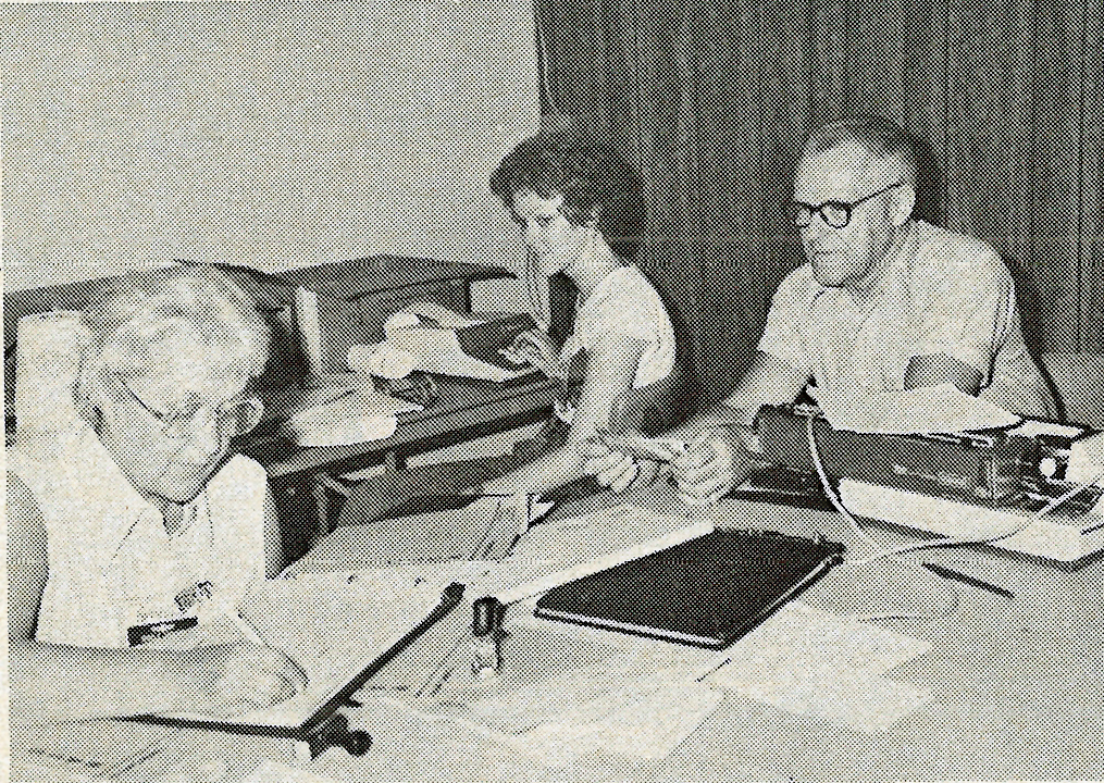 Three people working at a table with papers and a typewriter.
