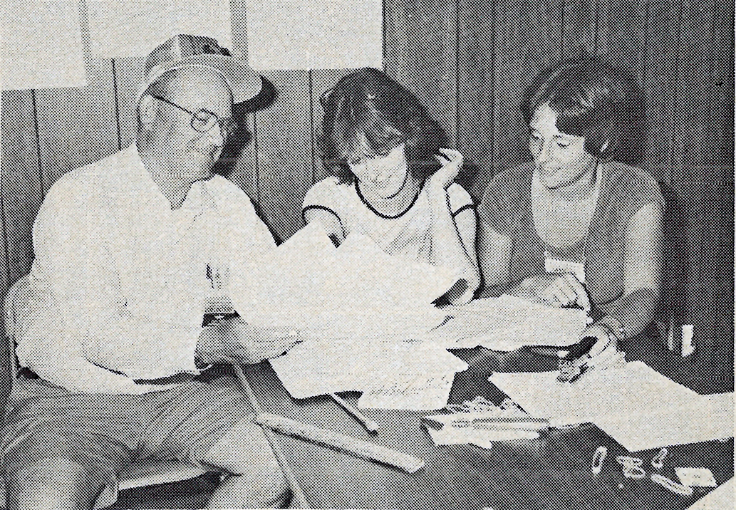 Three people at a table reviewing papers, two women and a man with a cap.