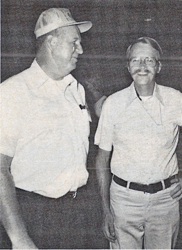 Two men smiling, one in a cap, both in casual shirts, black and white photo.