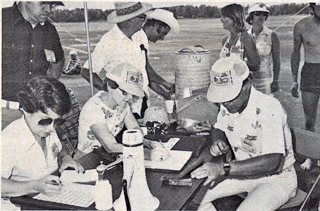 People in hats at a table, writing on papers outdoors.