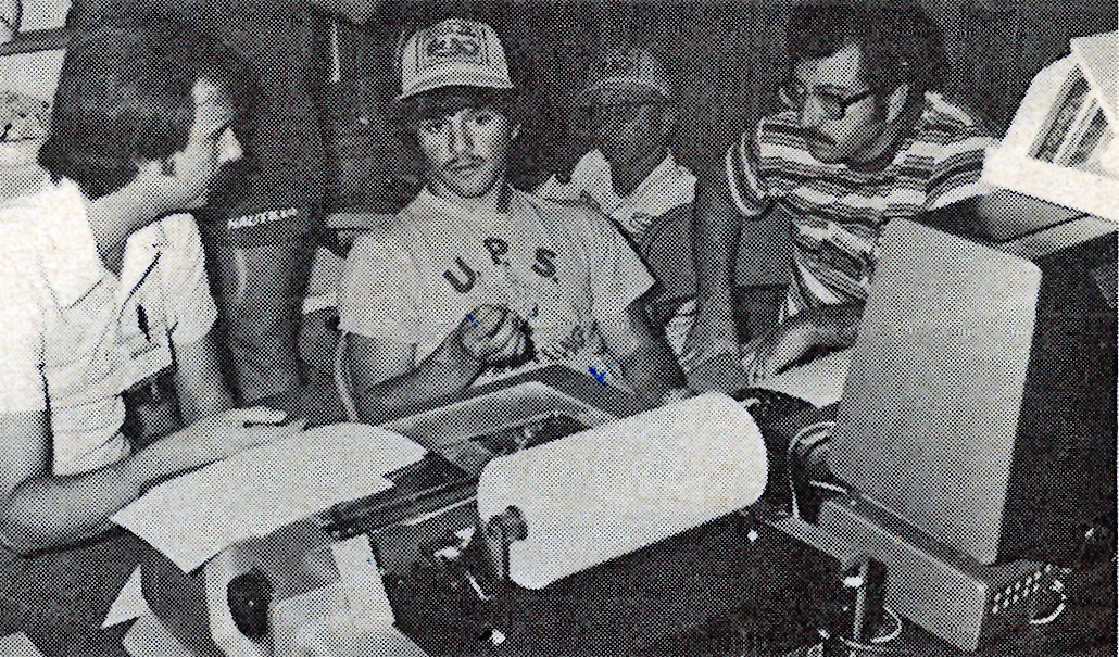 Black and white photo of four people discussing near a dot-matrix printer.