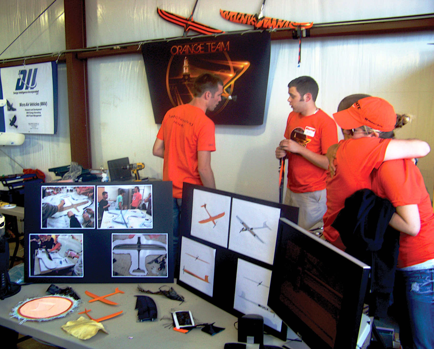 Booth with people in orange shirts, model planes, displays, and a "Orange Team" sign.