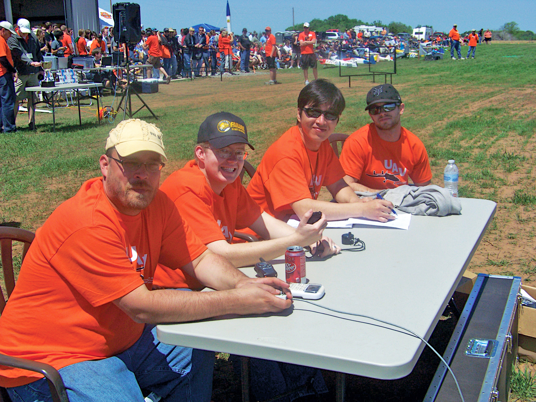Four men in orange shirts sit at an outdoor table, smiling at a crowded event.
