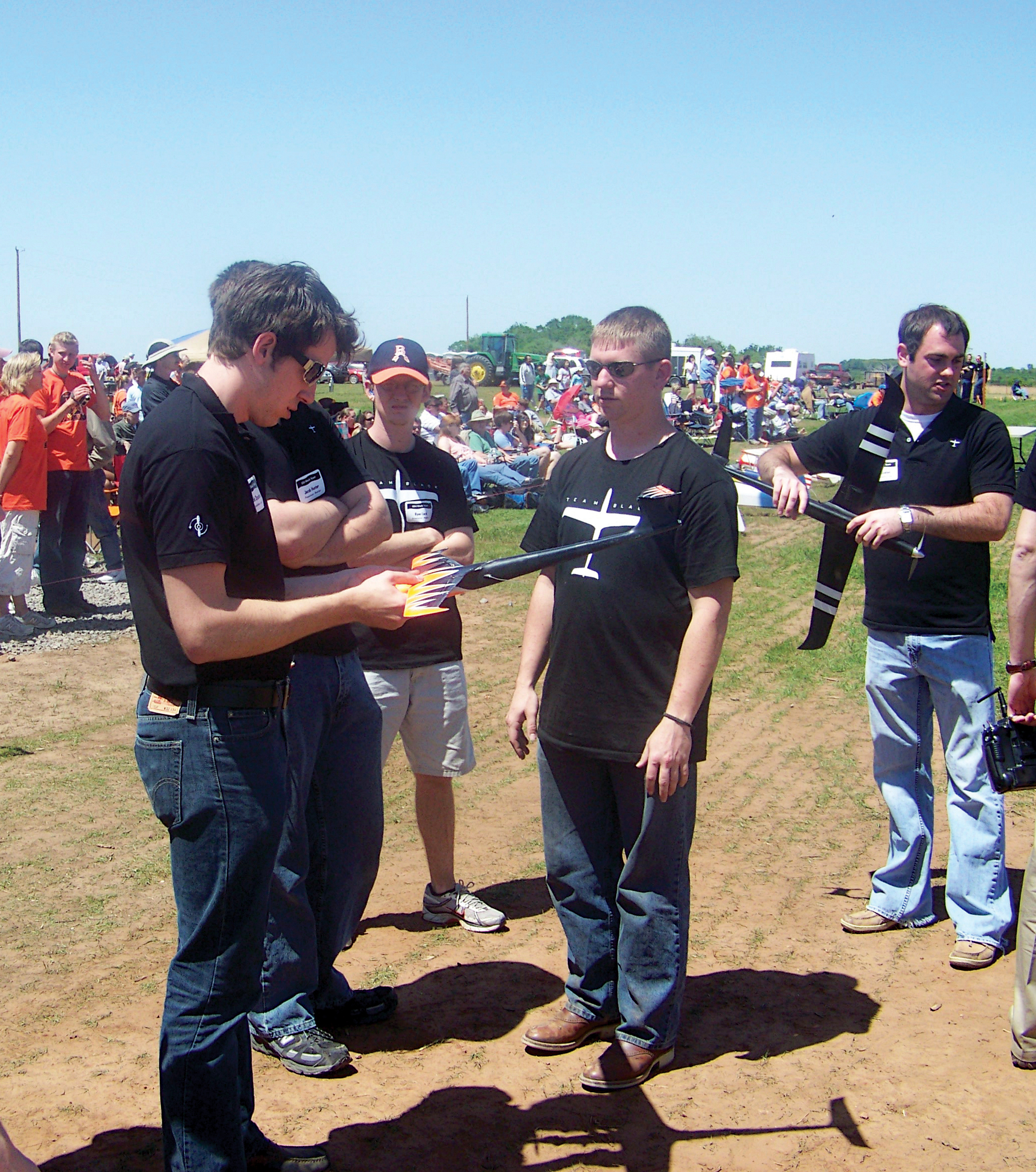 Men in black shirts discuss a model plane at an outdoor event on a sunny day.
