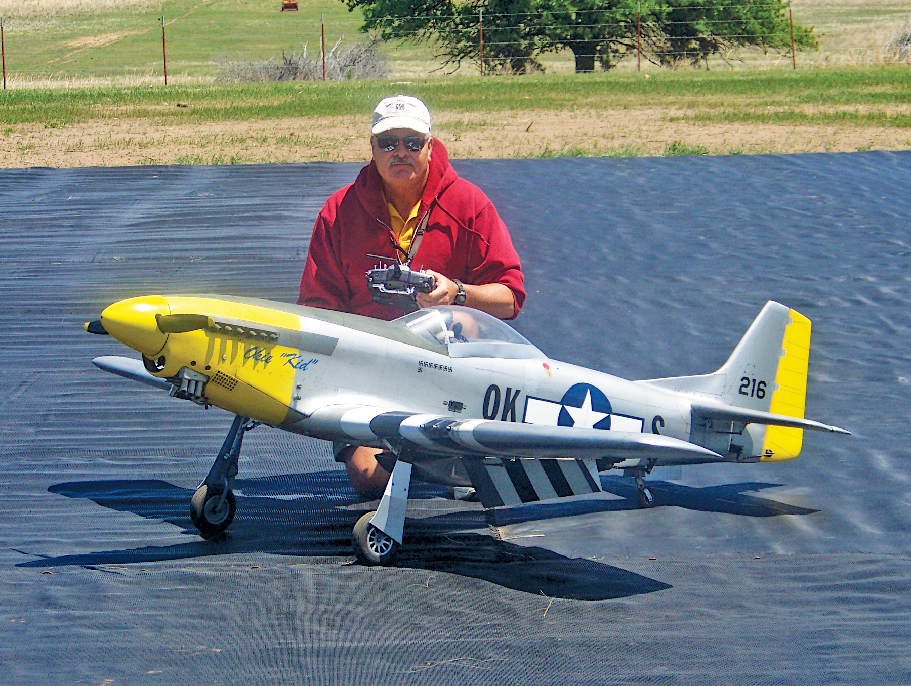 Man kneeling next to a large model airplane on a black mat outdoors.