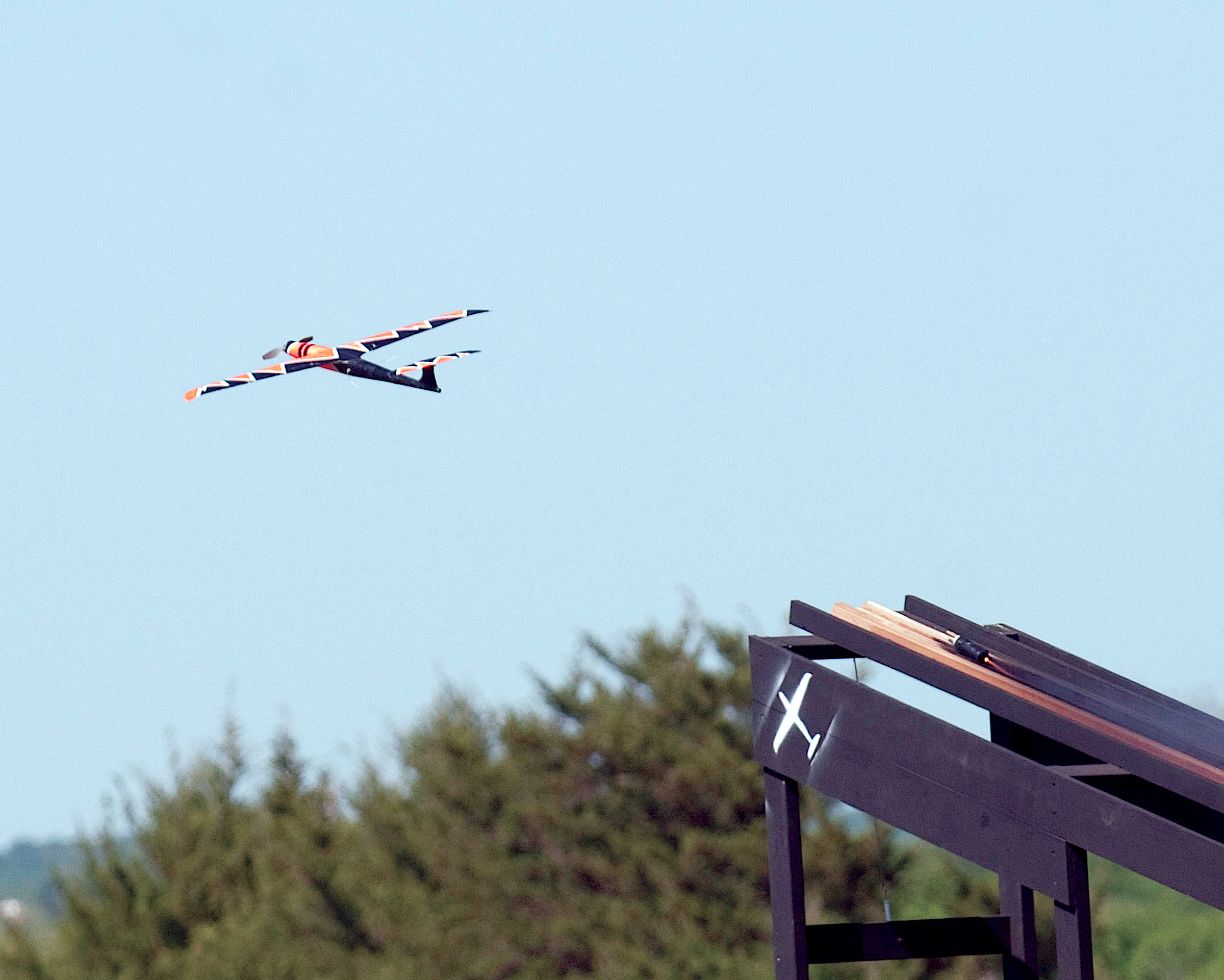 Small drone flying over trees and launching ramp in clear sky.