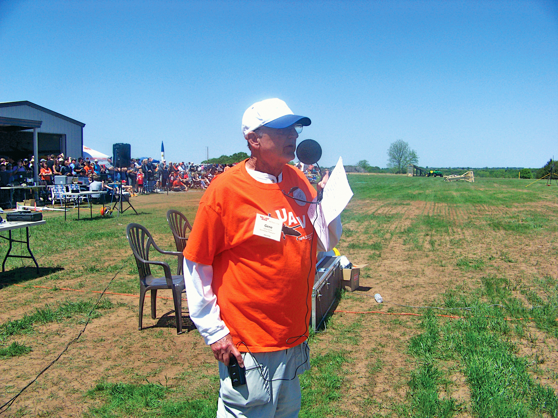 Man in an orange shirt holding a microphone outdoors, people gathered in the background.