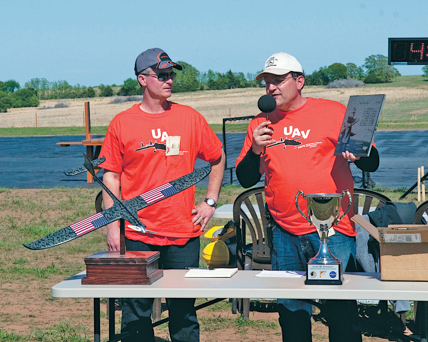 Two men in red shirts at an outdoor event with a trophy and model airplane.
