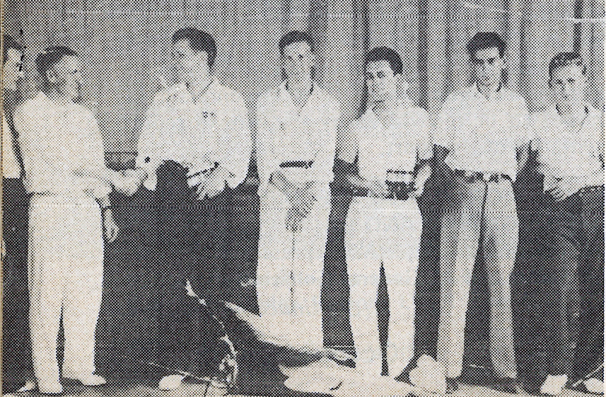 Black and white photo of seven men standing indoors, wearing casual clothing.