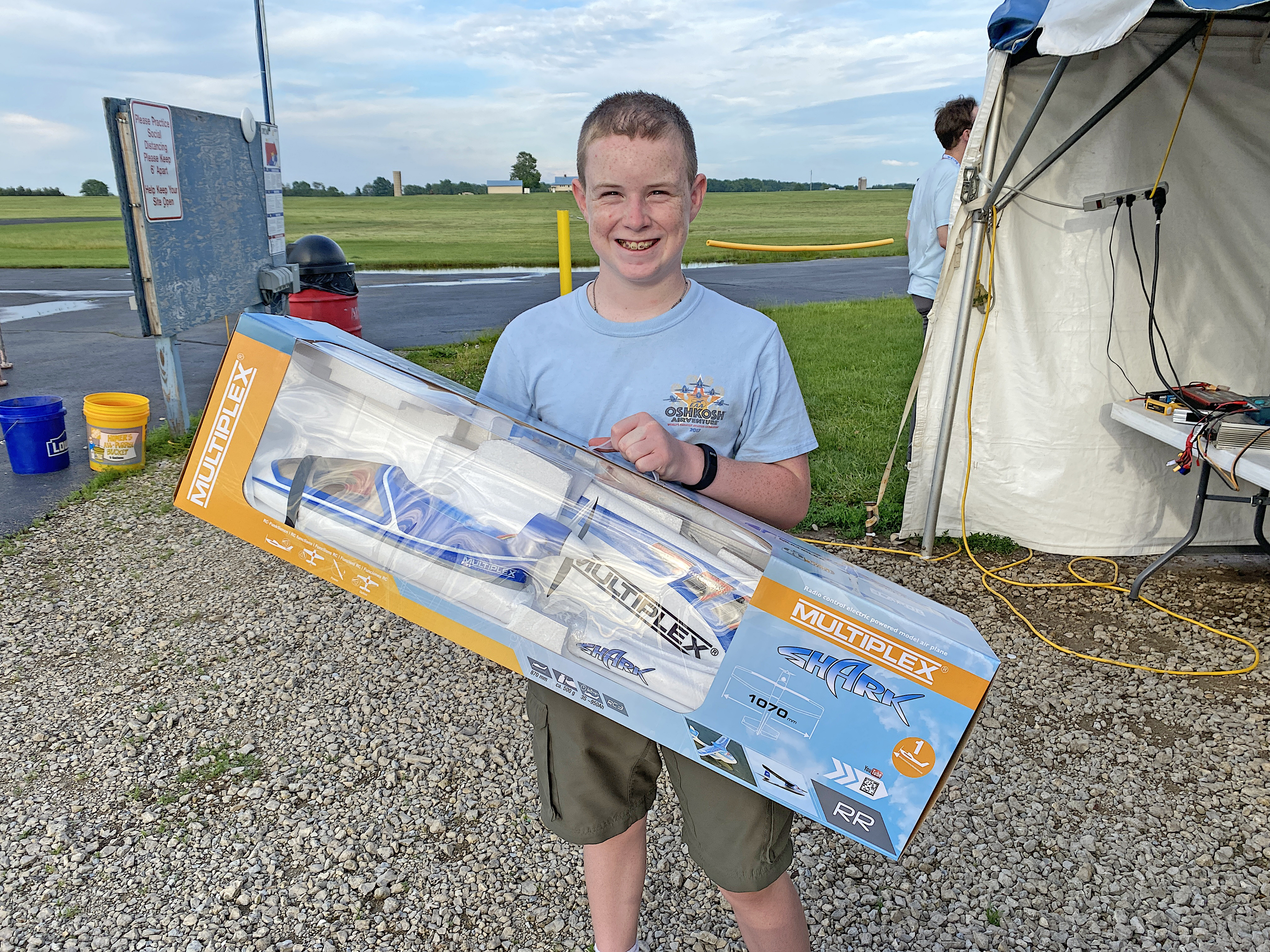 Teen boy smiles, holding a model plane box outdoors near a tent.