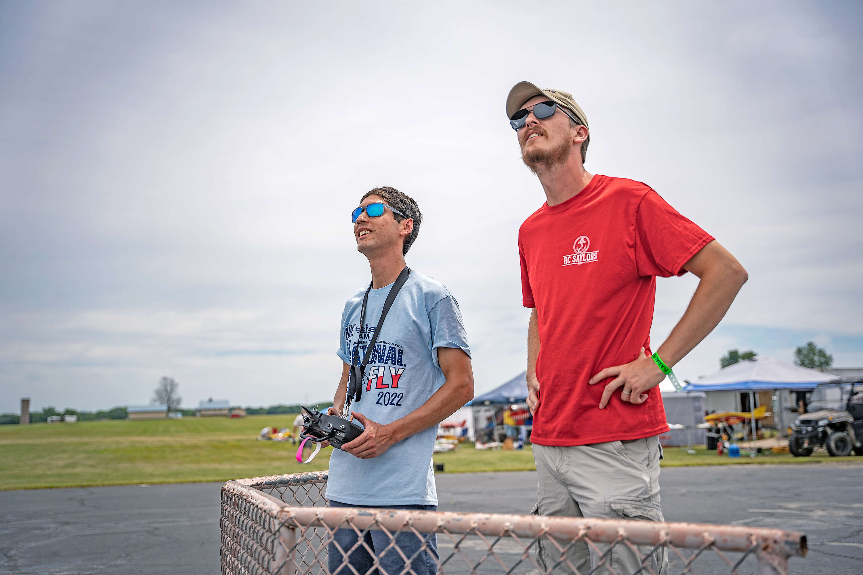 Two men wearing sunglasses and T-shirts watch the sky, holding a remote control.