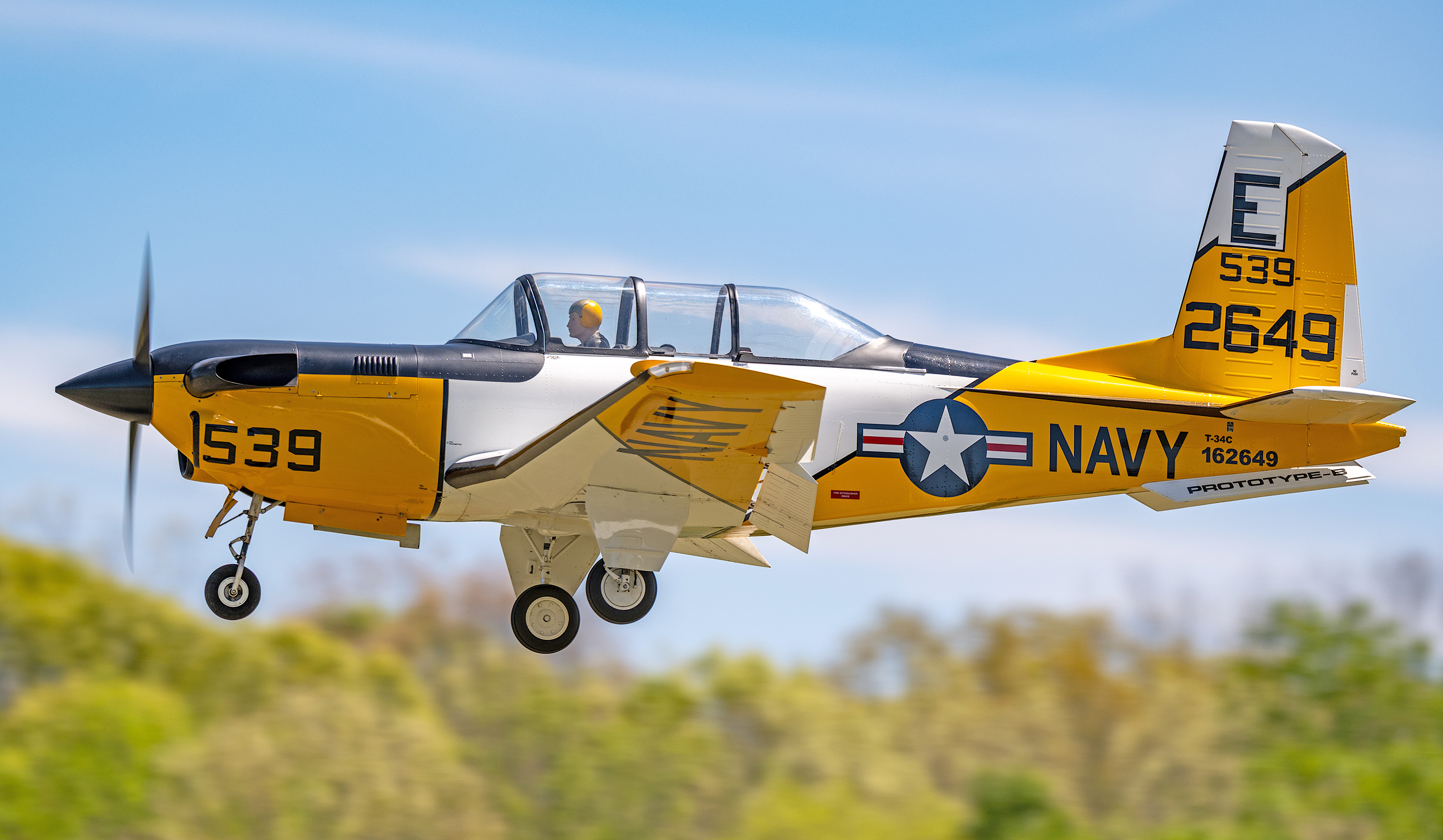 Navy aircraft with yellow and white design, flying against a blue sky.