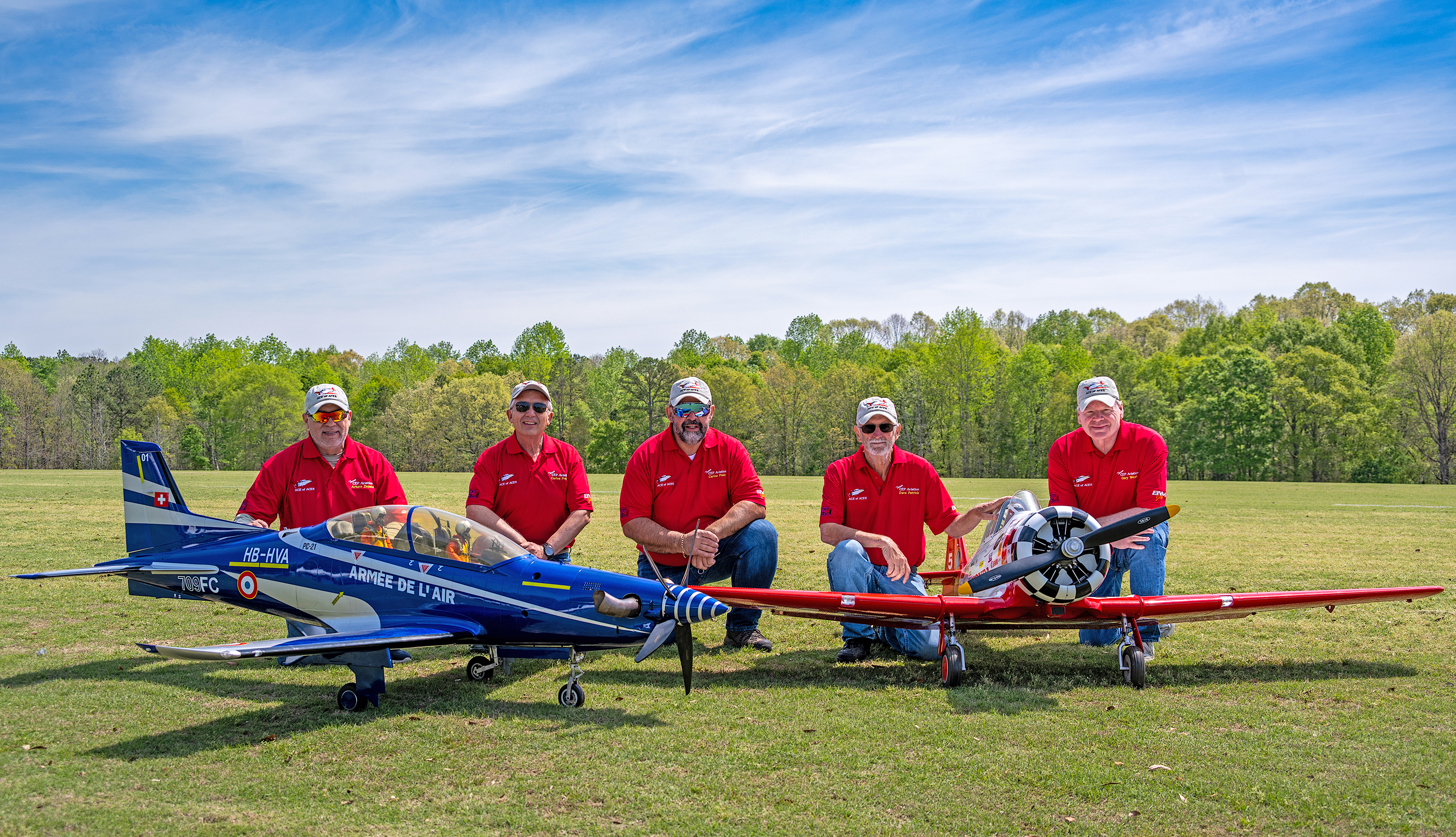 Five people in red shirts kneeling with model airplanes on a grassy field.