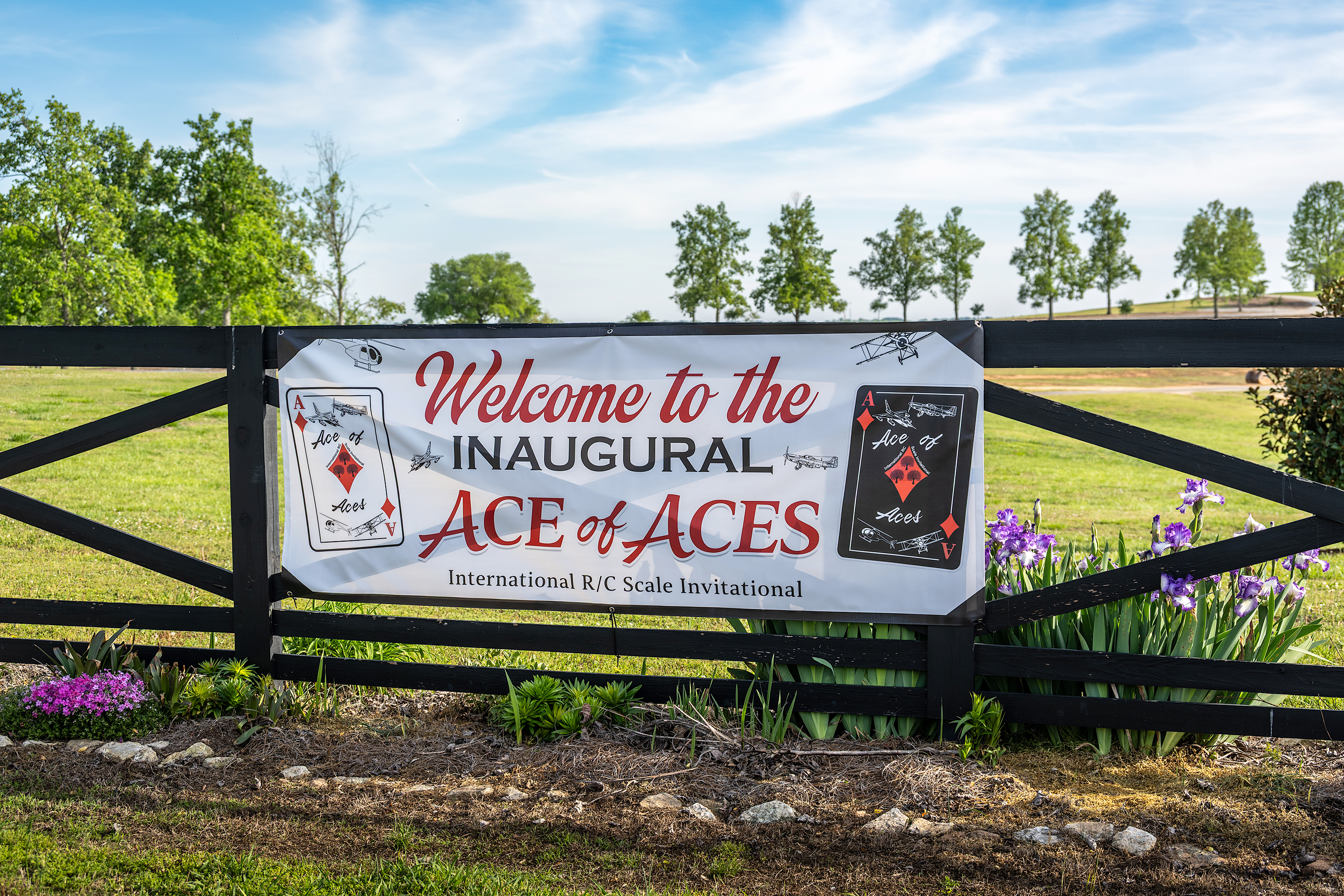 Welcome sign for the Inaugural Ace of Aces event on a black fence, with grass and flowers around.
