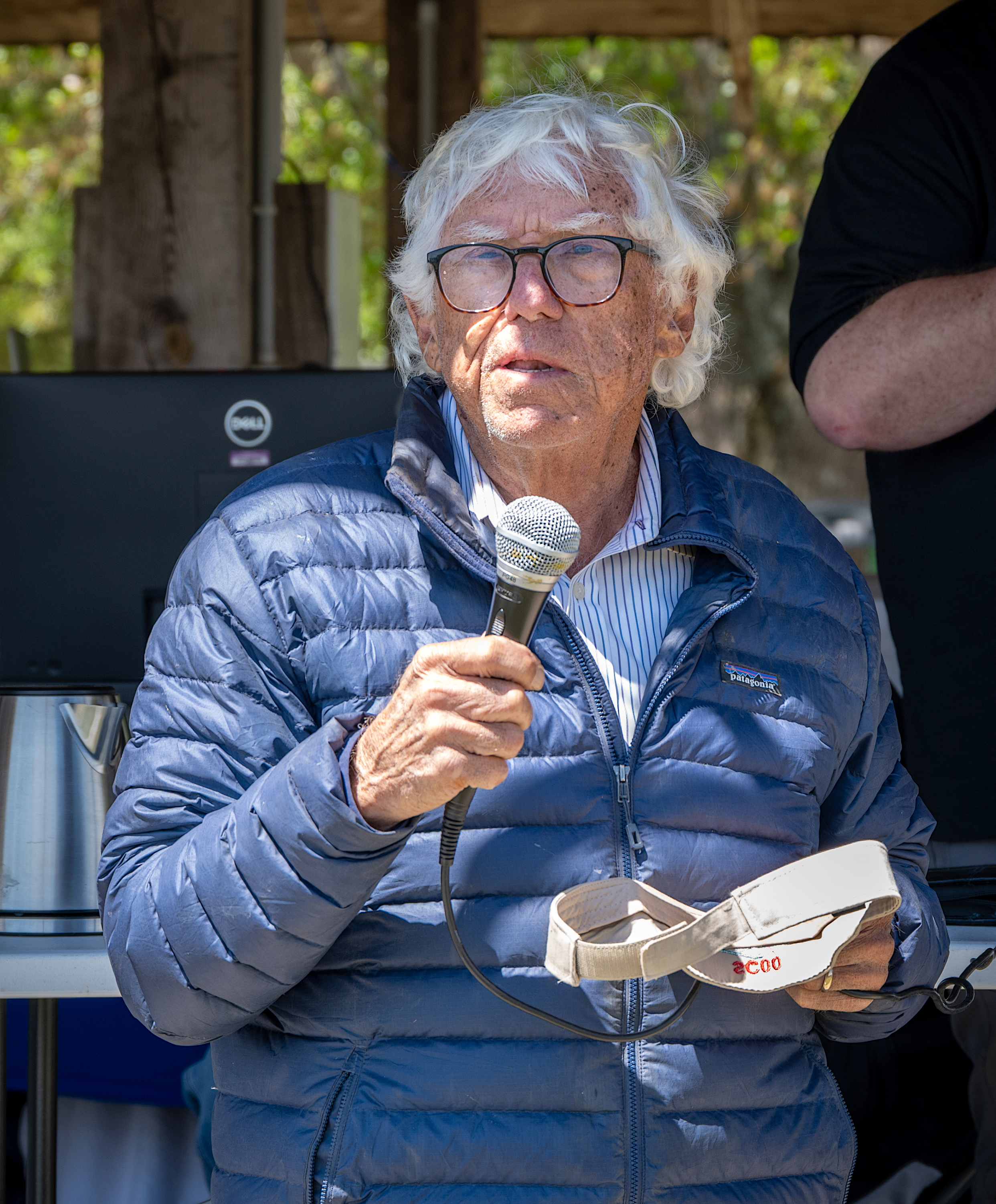Man with white hair and glasses speaking into a microphone outdoors.