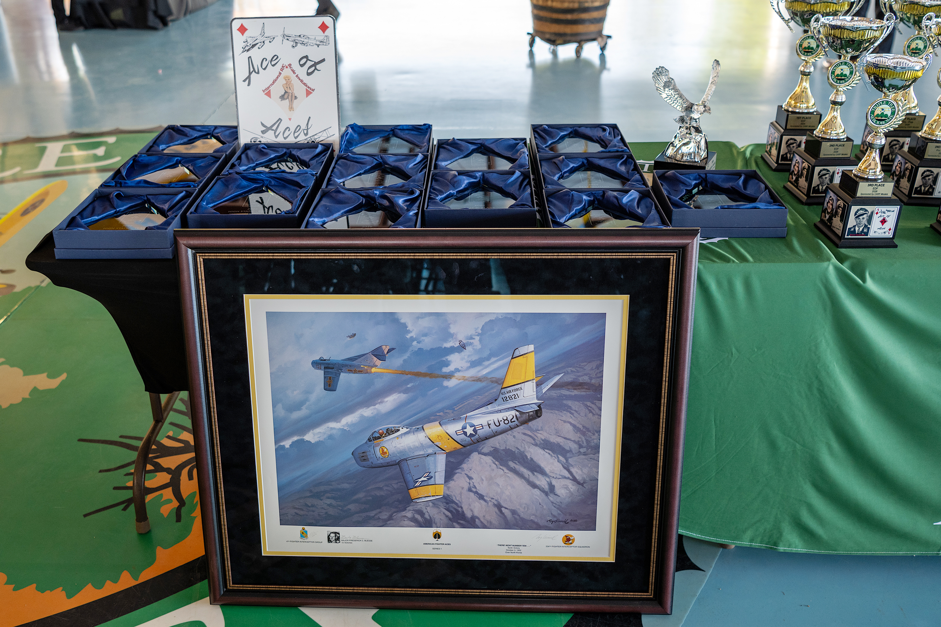 Framed airplane painting on table with awards and a "Let the Fuel Flow!" sign.