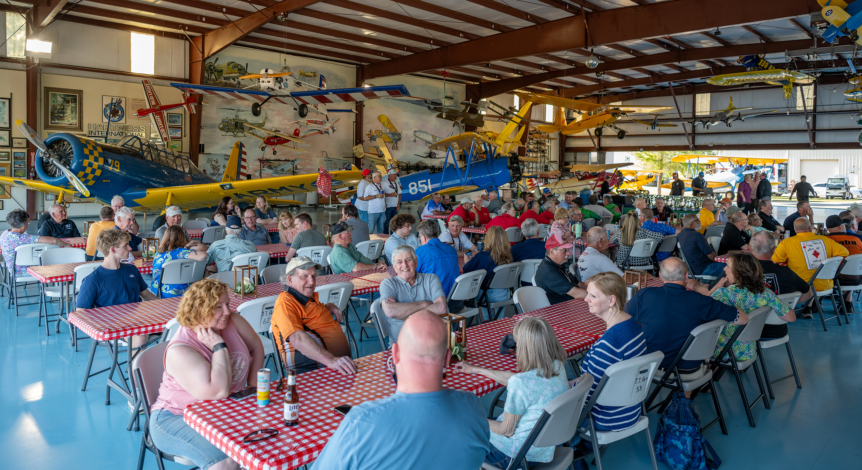 People seated at tables inside a hangar with vintage airplanes overhead.