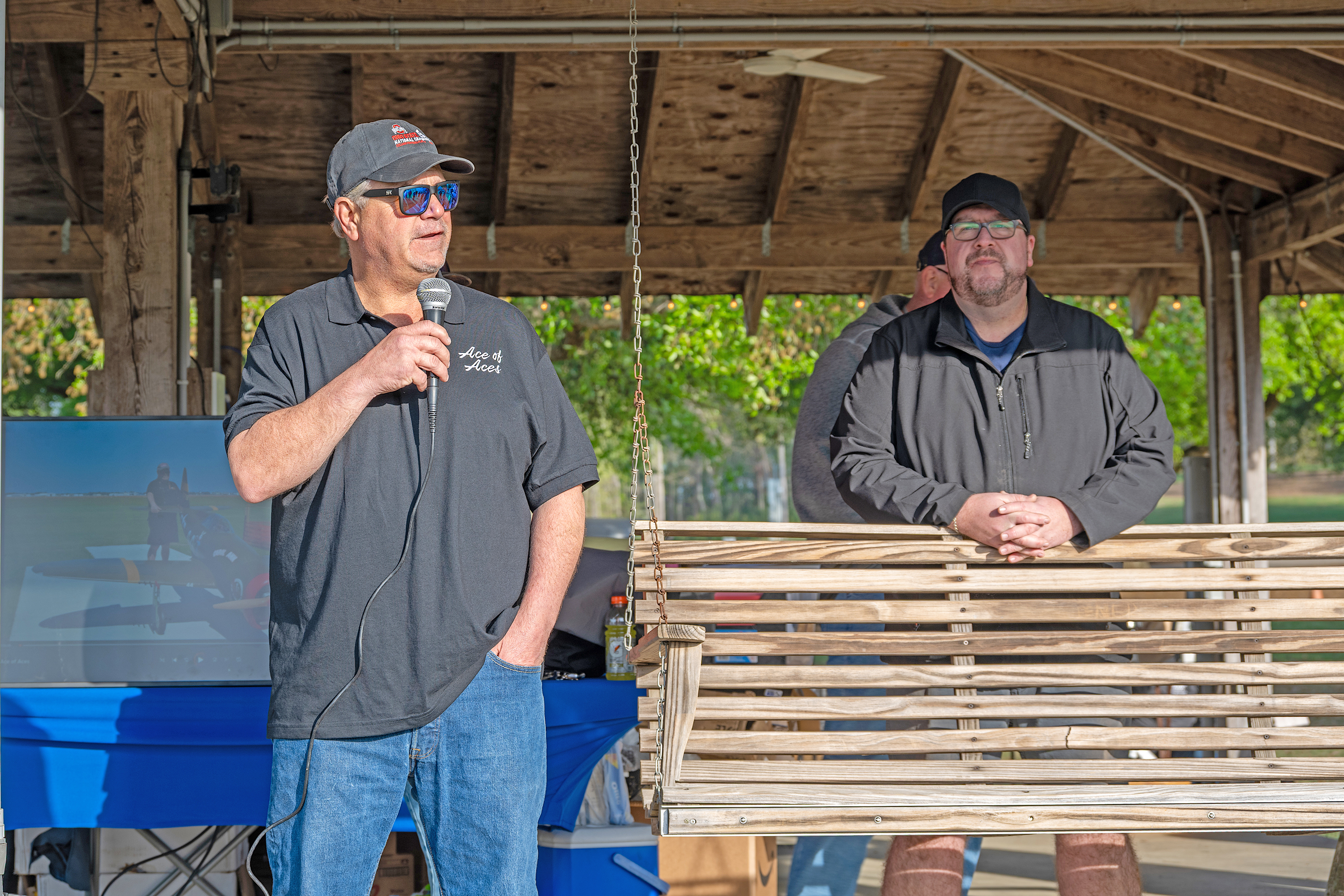 Man speaking into a microphone at an outdoor covered pavilion.
